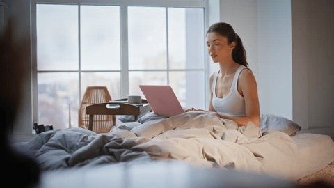 Vrouw leest op een laptop in bed bij het raam